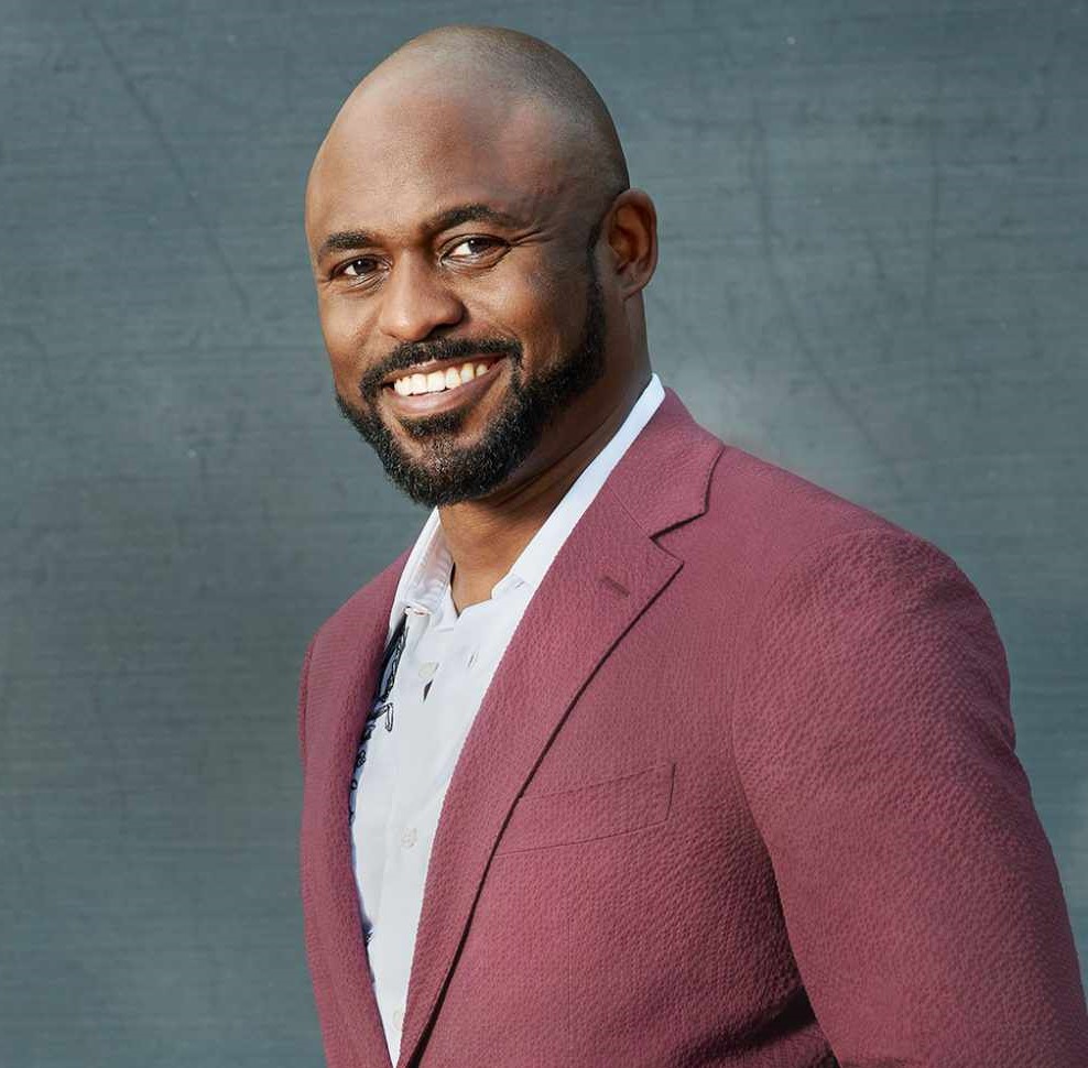 A publicity photo of Wayne Brady. A portrait of a man smiling at the camera wearing a magenta blazer