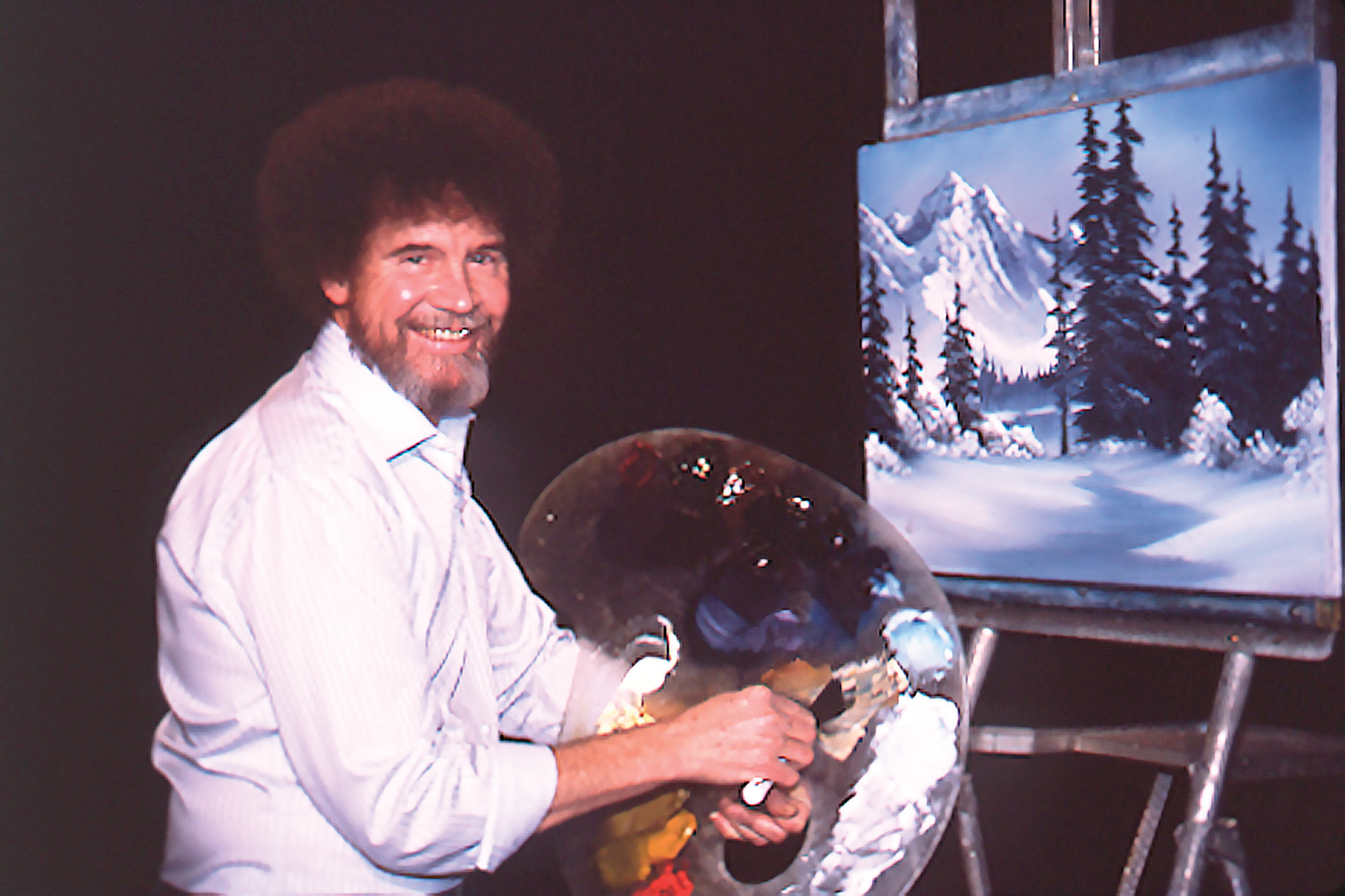 A photo of Bob Ross holding a painting palette in front of a painting of a winter mountain scene on the set of his television show